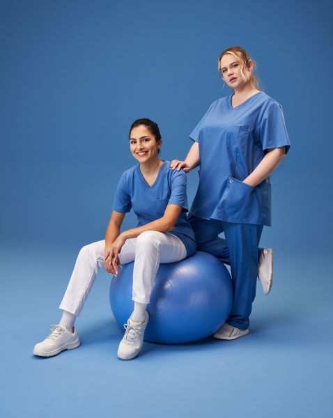Zwei Pflegerinnen tragen azurfarbene Arbeitskleidung. Eine davon sitzt  auf einem Gymnastikball.